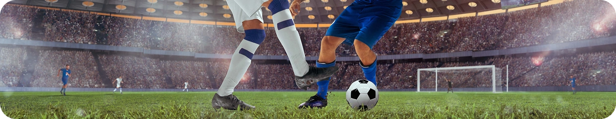 Gros plan sur les jambes de deux footballeurs et le ballon au niveau de la pelouse pendant un match, avec les tribunes du stade en arrière-plan (format bannière).