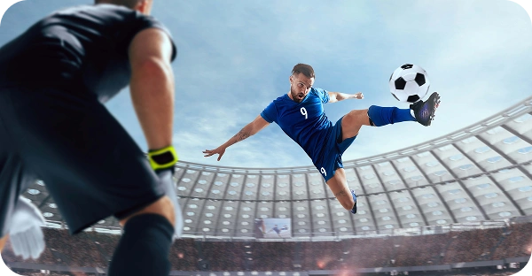 Un joueur de football en maillot bleu saute dans les airs pour frapper le ballon de volée devant le gardien de but dans un grand stade