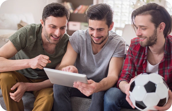 Trois amis assis sur un canapé regardent une tablette numérique, l'un d'eux tient un ballon de football