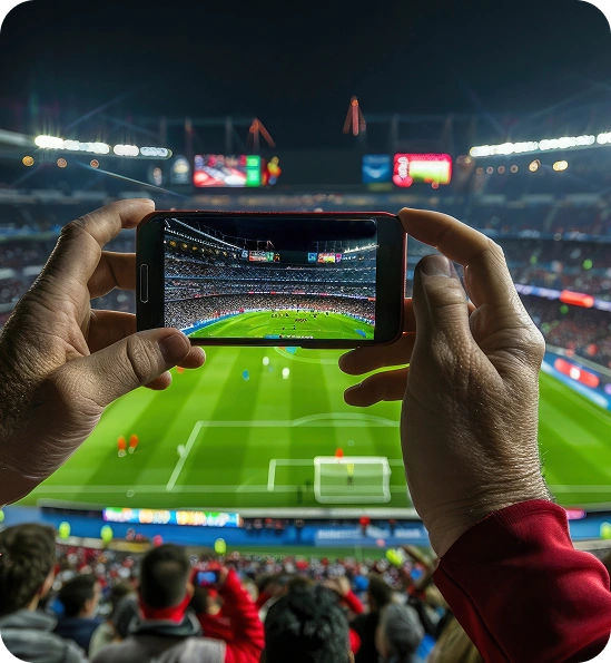 Mains d'un spectateur tenant un smartphone pour enregistrer ou photographier un match de football en direct depuis les tribunes d'un stade bondé.